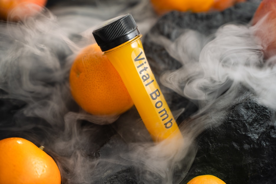 A test tube labeled 'Vital Bomb' is surrounded by oranges, with mist or fog creating a dramatic effect on a dark textured surface.