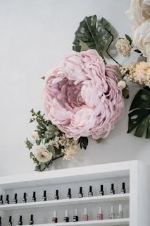 A large, soft pink flower arrangement is displayed on a wall, surrounded by green and white foliage. Below the floral display is a white shelf filled with various bottles of nail polish, arranged in rows.