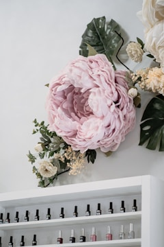 A large, soft pink flower arrangement is displayed on a wall, surrounded by green and white foliage. Below the floral display is a white shelf filled with various bottles of nail polish, arranged in rows.