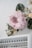 A large, soft pink flower arrangement is displayed on a wall, surrounded by green and white foliage. Below the floral display is a white shelf filled with various bottles of nail polish, arranged in rows.