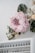 A large, soft pink flower arrangement is displayed on a wall, surrounded by green and white foliage. Below the floral display is a white shelf filled with various bottles of nail polish, arranged in rows.