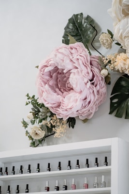 A large, soft pink flower arrangement is displayed on a wall, surrounded by green and white foliage. Below the floral display is a white shelf filled with various bottles of nail polish, arranged in rows.