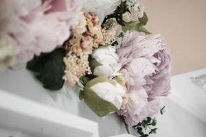 A bouquet of delicate flowers, featuring large pale pink roses, creamy white blooms, and clusters of blush carnations. The arrangement is complemented by green leaves and white petals, creating a soft, romantic aesthetic.