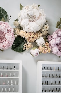 Large, soft-colored artificial flowers are mounted on a wall, creating a whimsical and elegant decor. Below the floral arrangement, two shelves display neatly organized nail polish bottles, each shelf having multiple rows filled with various colors.