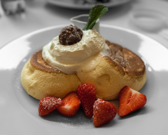 A plate with fluffy pancakes topped with a dollop of cream, a small amount of chocolate or caramel topping, and garnished with fresh strawberries and a mint leaf. The setting is light and airy, suggesting a relaxed breakfast or brunch scene.