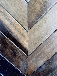 A detailed view of wooden planks arranged in a herringbone pattern. The wood showcases a variety of earthy tones, ranging from light to dark browns. The surface displays a natural texture with visible grains and small imperfections, adding to its rustic charm.