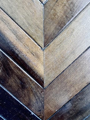 A detailed view of wooden planks arranged in a herringbone pattern. The wood showcases a variety of earthy tones, ranging from light to dark browns. The surface displays a natural texture with visible grains and small imperfections, adding to its rustic charm.