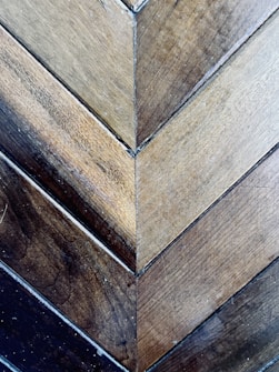 A detailed view of wooden planks arranged in a herringbone pattern. The wood showcases a variety of earthy tones, ranging from light to dark browns. The surface displays a natural texture with visible grains and small imperfections, adding to its rustic charm.