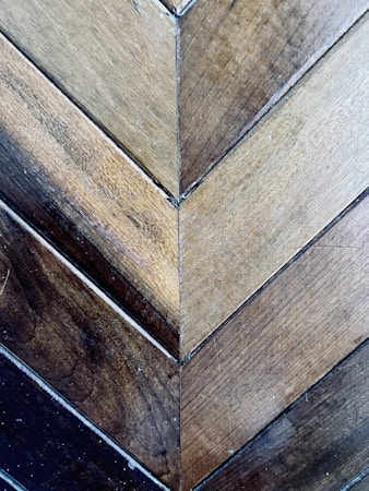A detailed view of wooden planks arranged in a herringbone pattern. The wood showcases a variety of earthy tones, ranging from light to dark browns. The surface displays a natural texture with visible grains and small imperfections, adding to its rustic charm.