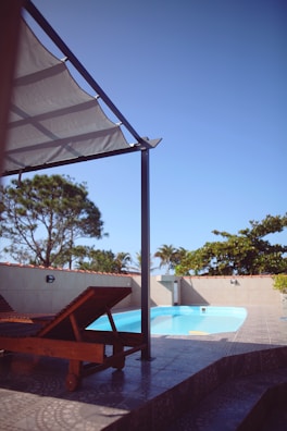 A serene outdoor scene featuring a swimming pool, surrounded by a light-colored tiled patio. Two wooden lounge chairs are placed under a modern pergola. In the background, trees with green foliage are silhouetted against a clear blue sky.