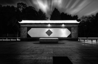 A nighttime scene depicting a structure with traditional architectural elements, featuring a symmetric design highlighted by decorative patterns. The sky shows a dynamic motion blur effect, creating a surreal ambiance. Trees line the background, silhouetted against the illuminated structure.