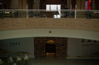 An interior architectural space features a two-level structure with a man standing on the upper level, which is adorned with detailed carvings of horse-drawn carriages. The lower level hosts floral arrangements, a stone wall entrance, and signage related to 'Tea Horse Road' in multiple languages.