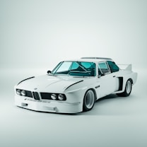 A classic white sports car with sleek aerodynamic features, prominent headlights, and a distinctive grille. The car is parked in a well-lit studio setting with a clean, minimal background. The design includes prominent wheel arches and a rear spoiler.