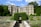 A grand historic mansion with a stone exterior and large windows is surrounded by lush greenery and manicured gardens. The pathway leading to the entrance is flanked by a stone railing and vibrant flower beds. The sky is bright with scattered clouds, enhancing the serene atmosphere.