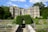 A grand historic mansion with a stone exterior and large windows is surrounded by lush greenery and manicured gardens. The pathway leading to the entrance is flanked by a stone railing and vibrant flower beds. The sky is bright with scattered clouds, enhancing the serene atmosphere.