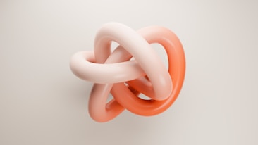 A visually striking intertwined structure with two tubular, smooth, and shiny loops. The loops are colored in shades of soft pink and coral, presenting an abstract geometric form suspended on a plain light background.