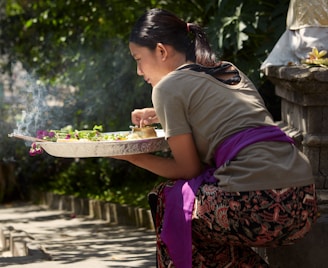 A person is kneeling outdoors near a stone structure, holding a tray with offerings possibly for a ritual. The tray contains green leaves and vibrant flowers with some incense smoke curling upwards. The individual wears patterned clothing with a purple sash and has neatly tied back hair. Sunlight filters through trees, casting dappled shadows on the ground.