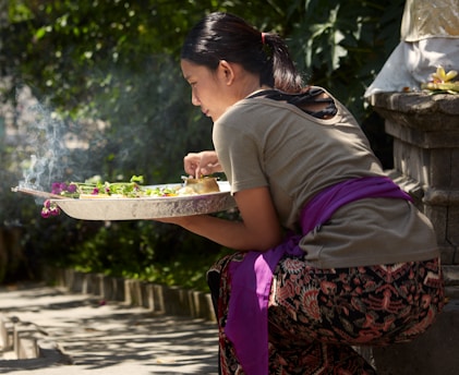 A person is kneeling outdoors near a stone structure, holding a tray with offerings possibly for a ritual. The tray contains green leaves and vibrant flowers with some incense smoke curling upwards. The individual wears patterned clothing with a purple sash and has neatly tied back hair. Sunlight filters through trees, casting dappled shadows on the ground.