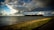 A large cargo ship sails across a vast body of water under dramatic, cloudy skies. The foreground features a grassy embankment bordering a narrow road, with patches of green visible in the shallow water.