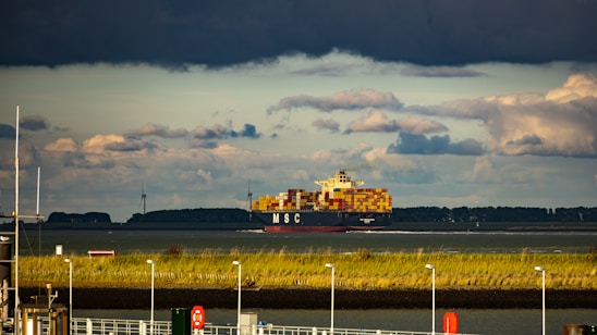 A cargo ship loaded with colorful containers is sailing across a body of water. The sky is filled with dark and dramatic clouds, creating a stark contrast with the lower horizon. Wind turbines are visible in the distance, and the shoreline is lined with grassy vegetation and several poles.