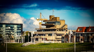 A concrete multi-story building with industrial architecture is seen amidst a mix of other buildings. A crane is positioned near the top of the building, and flags can be seen flying on poles. The sky is partly cloudy with a deep blue hue, and green grassy areas are visible in the foreground.