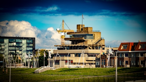 A concrete multi-story building with industrial architecture is seen amidst a mix of other buildings. A crane is positioned near the top of the building, and flags can be seen flying on poles. The sky is partly cloudy with a deep blue hue, and green grassy areas are visible in the foreground.
