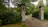 A wrought iron gate set between two brick pillars stands at the entrance of a cobblestone driveway. The area is surrounded by dense green foliage and various types of trees, creating a secluded and tranquil atmosphere.