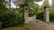 A wrought iron gate set between two brick pillars stands at the entrance of a cobblestone driveway. The area is surrounded by dense green foliage and various types of trees, creating a secluded and tranquil atmosphere.