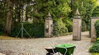 A cobblestone driveway leads to a set of ornate brick pillars topped with stone ornaments. Between the pillars, there is an open wrought iron gate leading to a pathway surrounded by lush green foliage. In the foreground, a green wheelbarrow with orange handles is placed on the driveway.