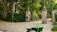 A cobblestone driveway leads to a set of ornate brick pillars topped with stone ornaments. Between the pillars, there is an open wrought iron gate leading to a pathway surrounded by lush green foliage. In the foreground, a green wheelbarrow with orange handles is placed on the driveway.