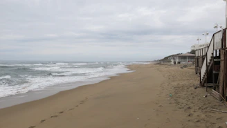 A deserted sandy beach stretches into the distance, with gentle waves lapping at the shore under a cloudy sky. A wooden stairway and building structures are visible on the right side, indicating a beachside establishment. The atmosphere appears calm and quiet, with footprints visible on the sand.