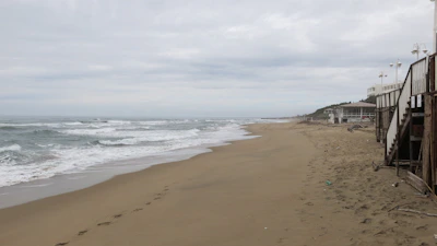 A deserted sandy beach stretches into the distance, with gentle waves lapping at the shore under a cloudy sky. A wooden stairway and building structures are visible on the right side, indicating a beachside establishment. The atmosphere appears calm and quiet, with footprints visible on the sand.