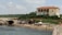 A scenic coastal area featuring a sandy beach with several people relaxing under umbrellas. To the left, there are limestone rock formations with arched entrances resembling small caves. An old, weathered building with faded paint and a red-tiled roof stands prominently in the background, surrounded by greenery.