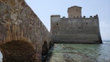 A stone bridge leads to a medieval fortress situated beside a body of water. The fortress features tall, thick walls with a square tower rising above. The walls show signs of age, with a texture of weathered stones and small windows. The surrounding water is calm with a hint of waves.