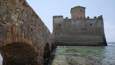 A stone bridge leads to a medieval fortress situated beside a body of water. The fortress features tall, thick walls with a square tower rising above. The walls show signs of age, with a texture of weathered stones and small windows. The surrounding water is calm with a hint of waves.
