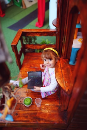A young child with a yellow headband and striped apron is seated on a wooden structure, holding a tablet displaying drawings. Beside the child is a green apple and a clear plastic cup. The image has vibrant and colorful surroundings, including a green carpet and colorful objects.
