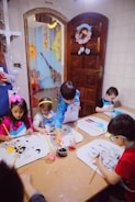 A group of young children are seated around a table, engaging in painting activities. Each child has a drawing in front of them, and they are using paintbrushes with containers of colorful paints. The room has cheerful decorations, and there is a wooden door with a floral wreath and light-colored tiles on the wall.
