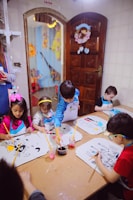 A group of young children are seated around a table, engaging in painting activities. Each child has a drawing in front of them, and they are using paintbrushes with containers of colorful paints. The room has cheerful decorations, and there is a wooden door with a floral wreath and light-colored tiles on the wall.