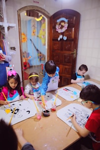 A group of young children are seated around a table, engaging in painting activities. Each child has a drawing in front of them, and they are using paintbrushes with containers of colorful paints. The room has cheerful decorations, and there is a wooden door with a floral wreath and light-colored tiles on the wall.