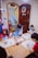 A group of young children are seated around a table, engaging in painting activities. Each child has a drawing in front of them, and they are using paintbrushes with containers of colorful paints. The room has cheerful decorations, and there is a wooden door with a floral wreath and light-colored tiles on the wall.