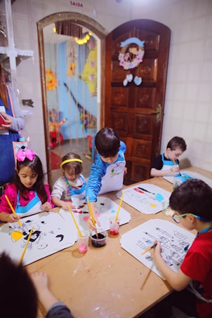 A group of young children are seated around a table, engaging in painting activities. Each child has a drawing in front of them, and they are using paintbrushes with containers of colorful paints. The room has cheerful decorations, and there is a wooden door with a floral wreath and light-colored tiles on the wall.