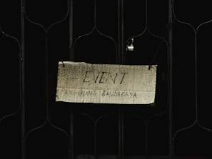 A handwritten sign on a piece of cardboard is hung on a metal gate. The sign reads 'EVENT' with additional text underneath that is partially visible. It appears to be suspended using wires or strings attached to the gate.