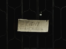A handwritten sign on a piece of cardboard is hung on a metal gate. The sign reads 'EVENT' with additional text underneath that is partially visible. It appears to be suspended using wires or strings attached to the gate.