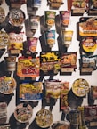 A variety of packaged instant noodle products are displayed on a wall, each with vibrant and colorful packaging. The items include cup noodles and packet noodles, showcasing diverse flavors and brands, primarily featuring Asian script and designs.