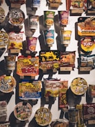 A variety of packaged instant noodle products are displayed on a wall, each with vibrant and colorful packaging. The items include cup noodles and packet noodles, showcasing diverse flavors and brands, primarily featuring Asian script and designs.