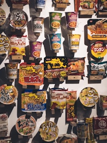 A variety of packaged instant noodle products are displayed on a wall, each with vibrant and colorful packaging. The items include cup noodles and packet noodles, showcasing diverse flavors and brands, primarily featuring Asian script and designs.