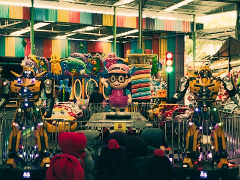 An indoor amusement area featuring colorful decorations with cartoonish designs. There are large robot-like figures positioned prominently alongside vibrant ride-on cars for children. Various colorful designs cover the walls and ceilings, reflecting a playful and fantasy-themed setting.