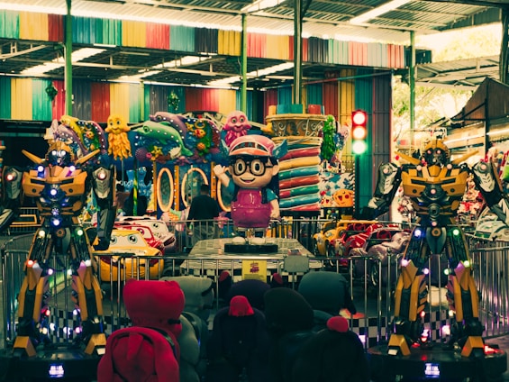 An indoor amusement area featuring colorful decorations with cartoonish designs. There are large robot-like figures positioned prominently alongside vibrant ride-on cars for children. Various colorful designs cover the walls and ceilings, reflecting a playful and fantasy-themed setting.