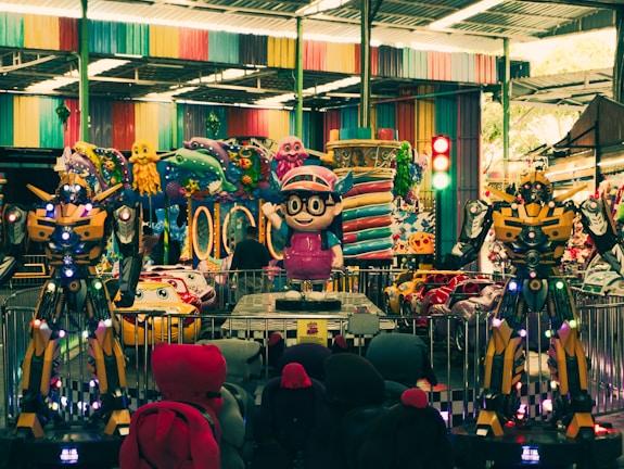 An indoor amusement area featuring colorful decorations with cartoonish designs. There are large robot-like figures positioned prominently alongside vibrant ride-on cars for children. Various colorful designs cover the walls and ceilings, reflecting a playful and fantasy-themed setting.