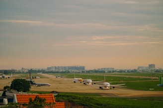 Multiple airplanes are queued on a runway at an airport, surrounded by green grass fields and bordered by a cityscape in the background under a cloudy sky.